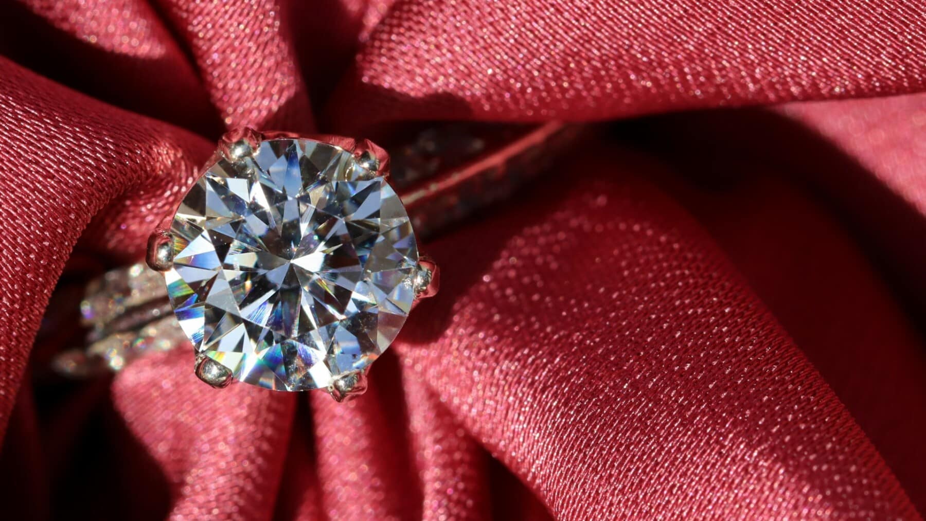 Close-up of a brilliant solitaire diamond engagement ring on luxurious red silk. The perfect ring for a romantic holiday proposal by Harry Glinberg Jewelers.